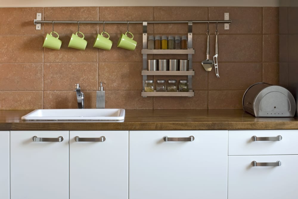 A kitchen with white base cupboard, brown counter and hooks mounted on the wall to hold mugs, spices and cheese cutting knives. 